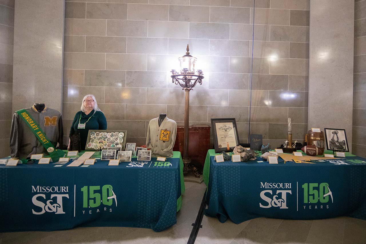 Woman stands behind table with 150 branded cover and historical items displayed.