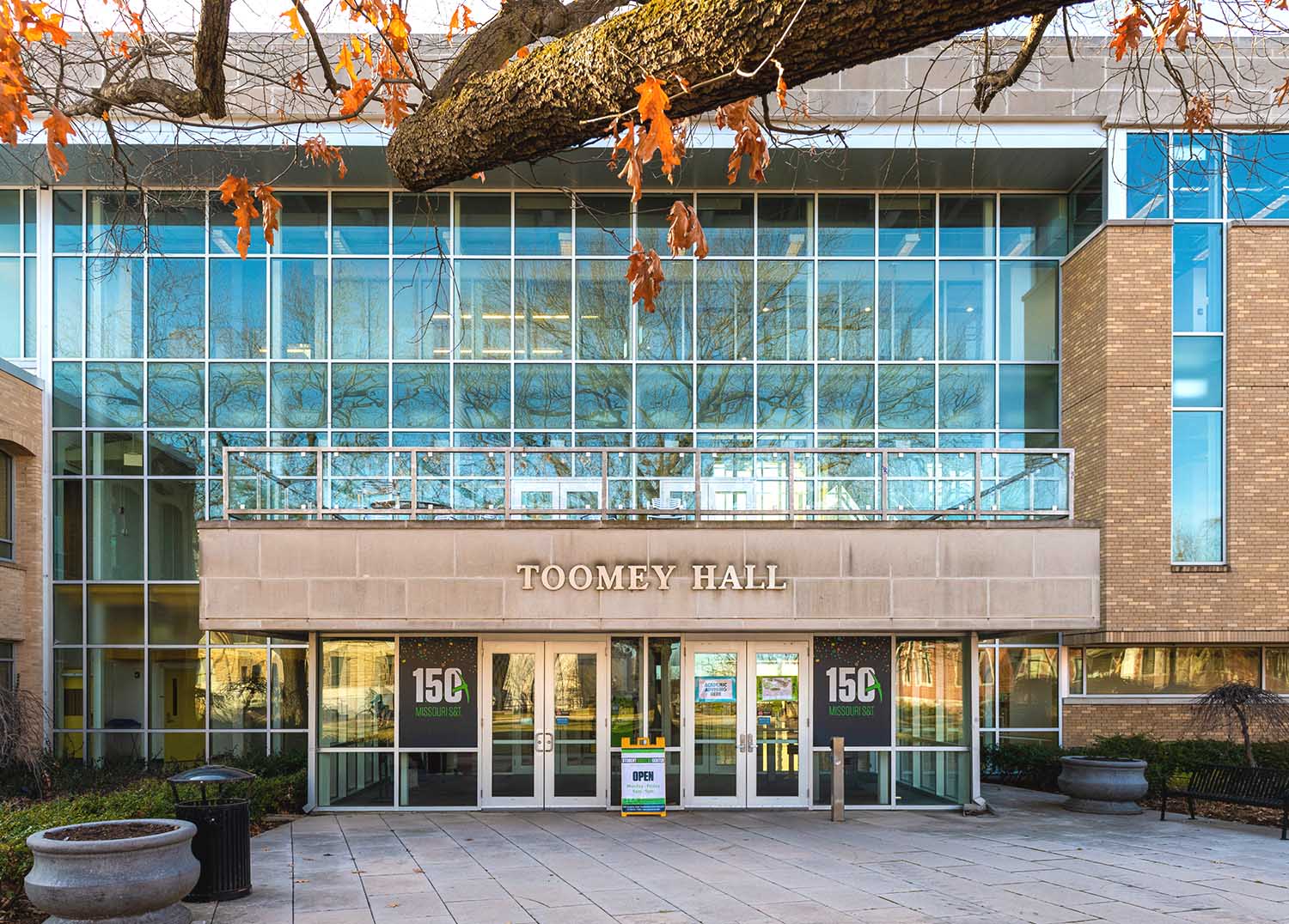 Front of building with glass windows and doors. 150 branded decals on the left and right of doors. Tree branch with orange leafs at the top.